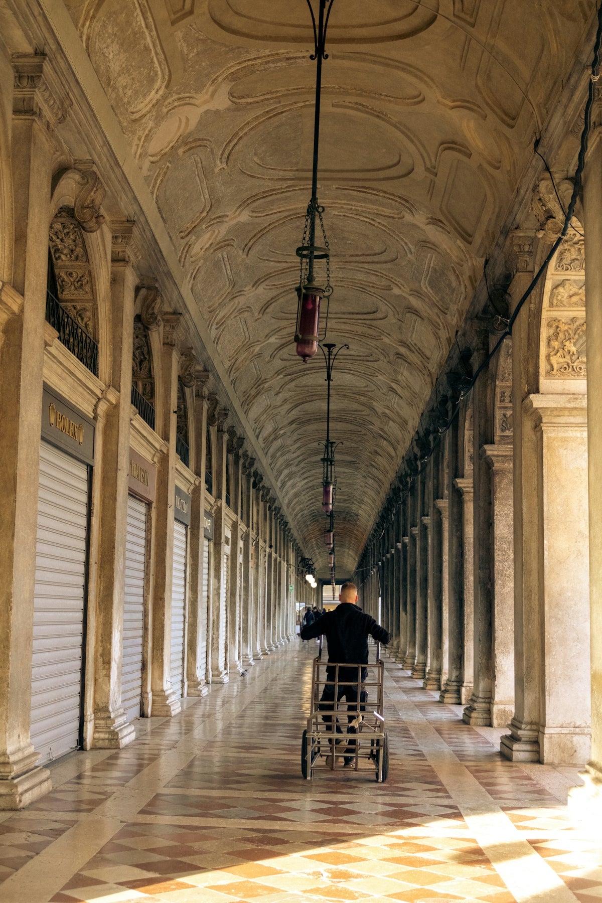 Person pushing a cart in a long, ornate arched arcade.